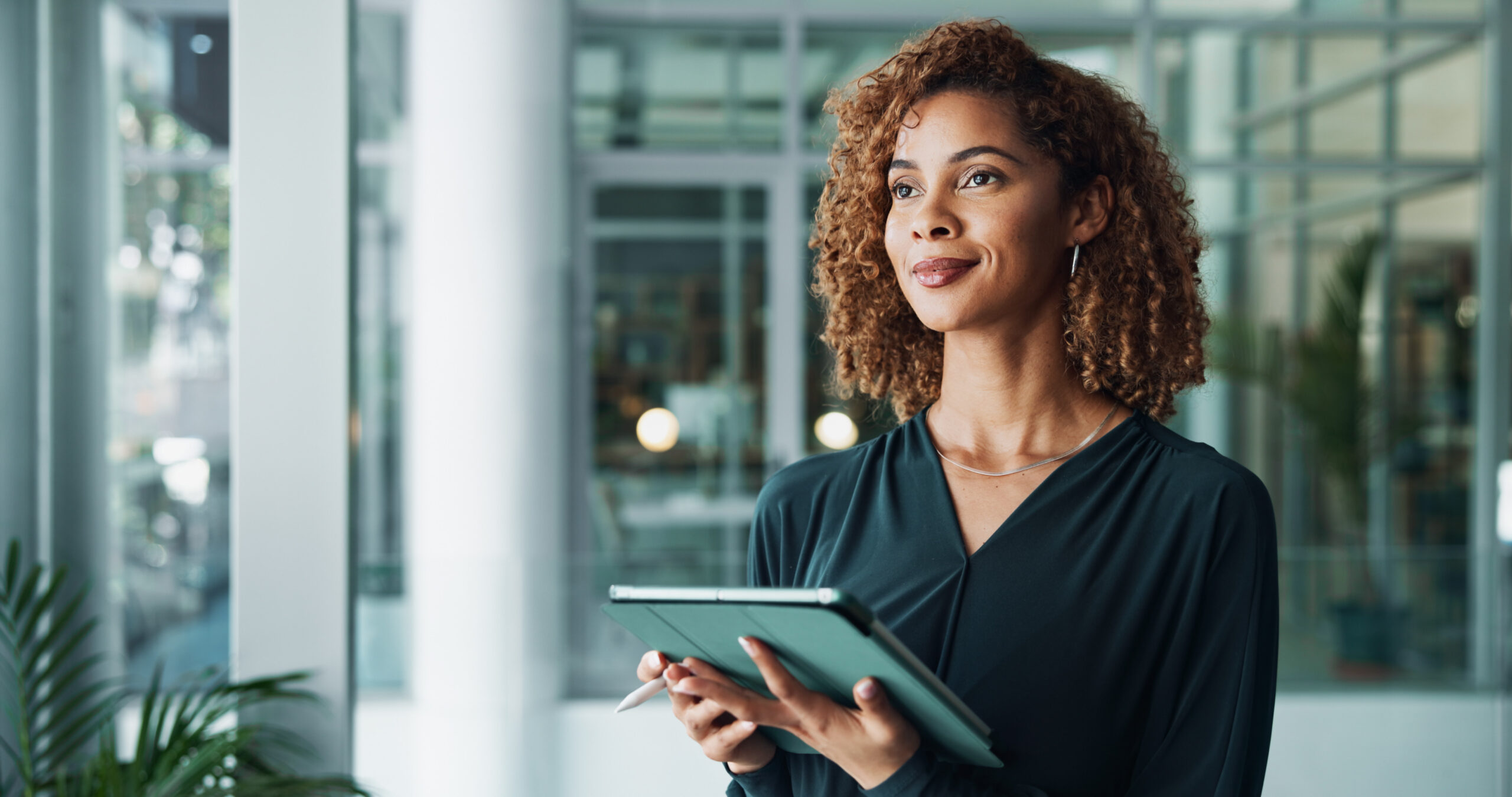Professional woman standing in a modern office holding a tablet and stylus, looking confidently into the distance.