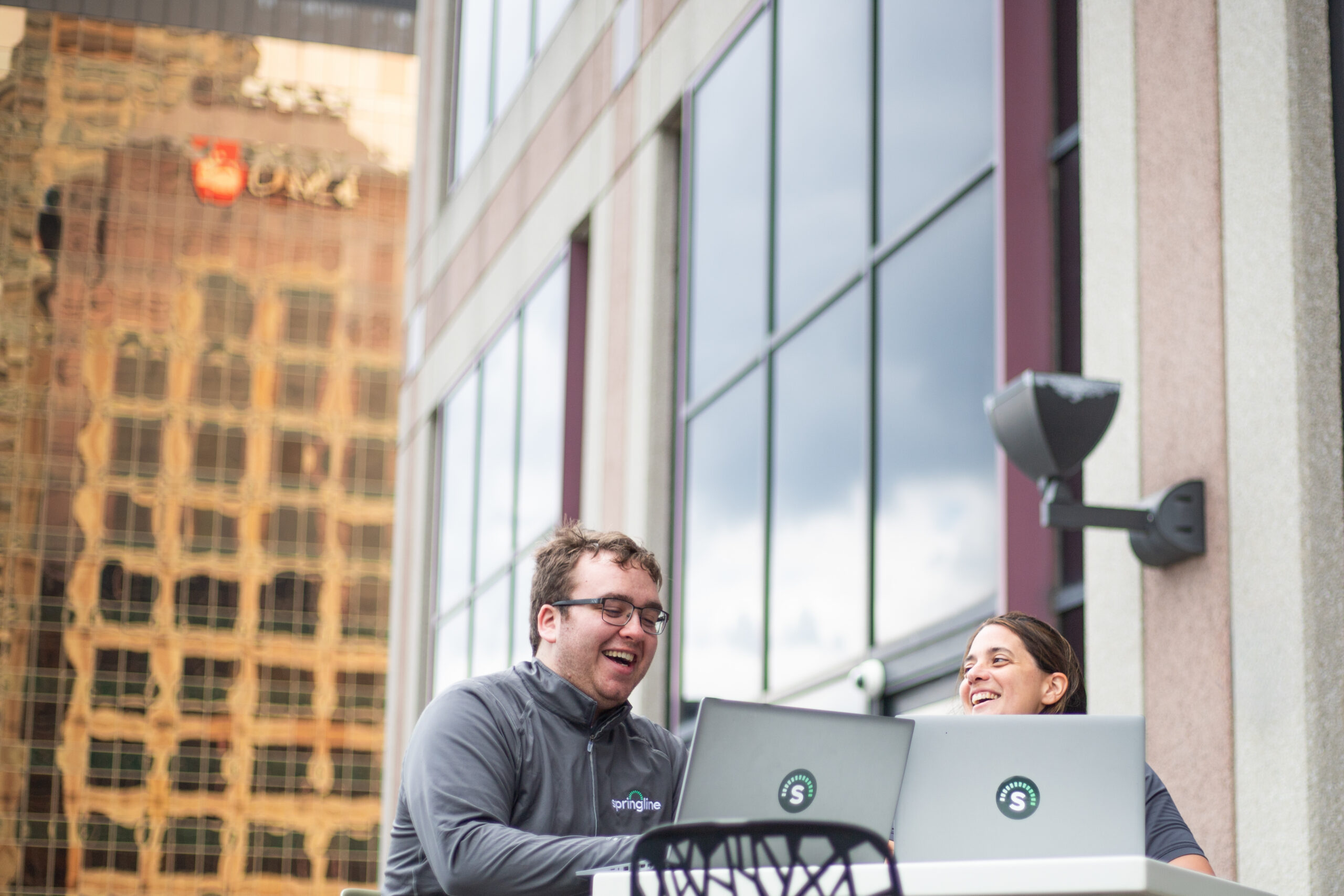 Two employees work together outdoors at a high-top table on a downtown office terrace, each using a Springline-branded laptop. They smile and laugh while collaborating, with modern office buildings and reflective glass windows creating an urban backdrop. The image highlights a flexible, connected work environment that extends beyond the traditional office.