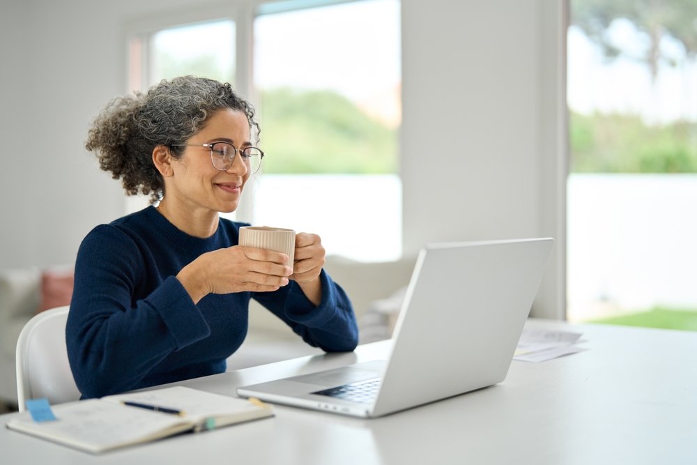 Image of a business professional smiling as she looks at her laptop, with a cup of coffee/tea in her hand.
